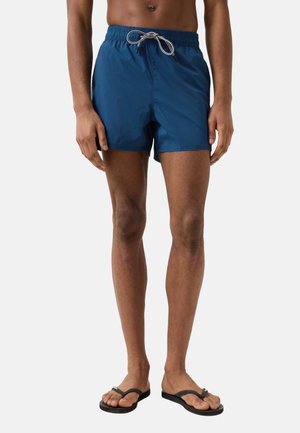 Mand iført marineblå badeshorts med hvid snor og sorte flip-flops, stående mod en ensartet lys baggrund.