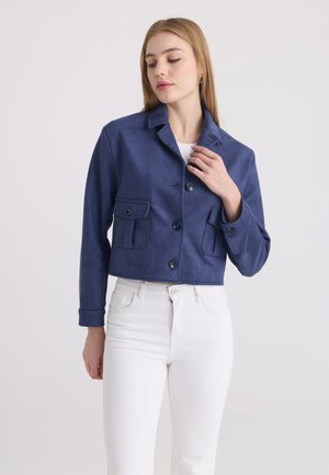 Femme portant une veste courte bleu marine avec des poches à rabats boutonnées et un pantalon blanc taille haute, debout devant un fond clair uni.