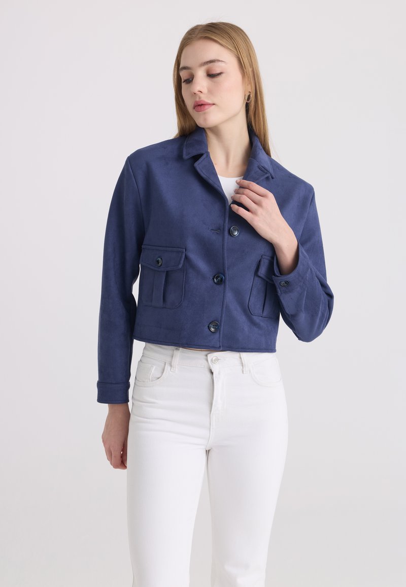 Femme portant une veste courte bleu marine avec des poches à rabats boutonnées et un pantalon blanc taille haute, debout devant un fond clair uni.