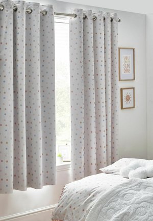 Tende bianche a pois pastello appese accanto a una finestra in una camera da letto dolcemente illuminata con biancheria coordinata e quadri incorniciati.