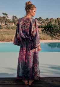 Kimono lung, diafan, în nuanțe bogate de violet și teal, cu model paisley. Are mâneci largi și talie strânsă, purtat peste o ținută simplă.