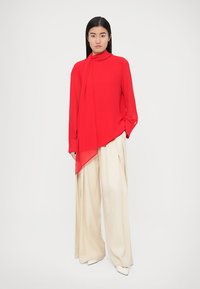 Blouse rouge à manches longues avec un devant drapé et un col montant, associée à un pantalon large beige, présentant une texture lisse et une légère brillance.