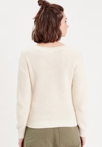 Cream knit sweater with a relaxed fit, featuring a textured weave, ribbed cuffs, and a wide neckline. Worn over olive green pants.