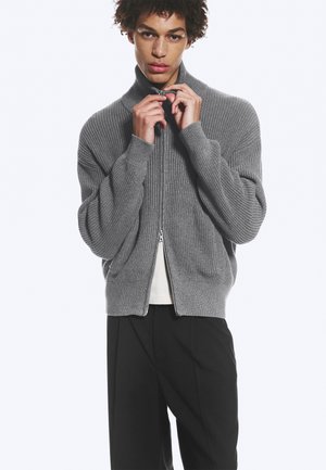 Joven cerrándose una chaqueta gris de punto acanalado, lleva pantalones negros y una camisa blanca debajo, sobre un fondo blanco liso.