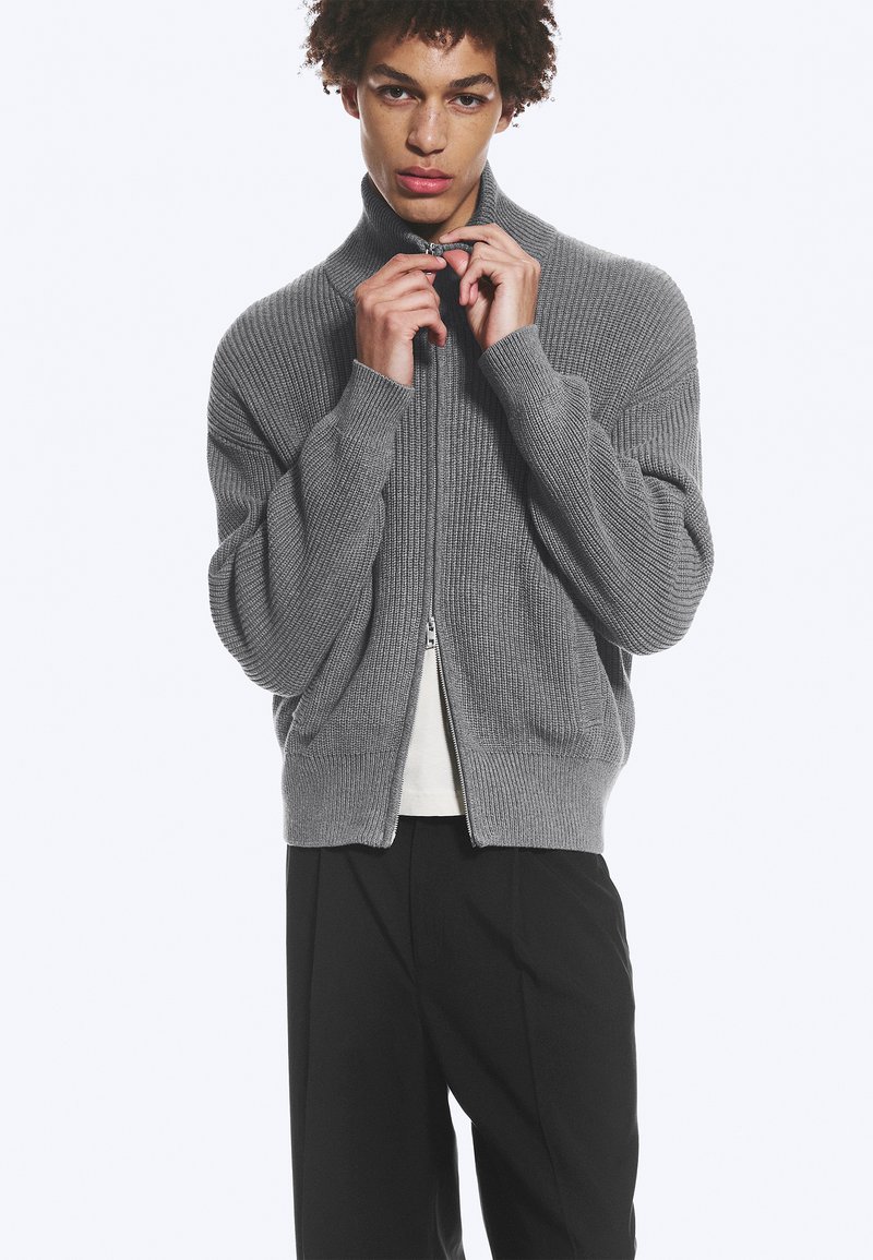 Jeune homme en train de zipper une veste en maille côtelée grise, portant un pantalon noir et une chemise blanche en dessous, sur un fond blanc uni.