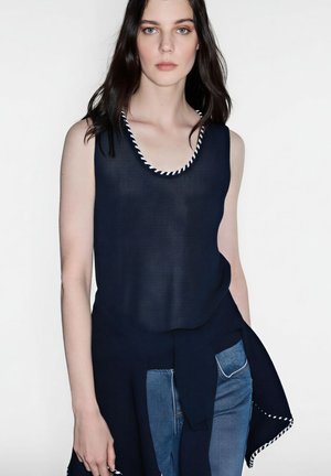 Mujer con cabello largo y oscuro, vestida con una camiseta sin mangas de punto azul marino con ribete blanco, sosteniendo un suéter azul marino atado a la cintura sobre unos vaqueros azules.