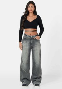 Top corto negro acanalado con mangas largas combinado con jeans de tiro alto y pierna ancha en gris claro; la modelo está de pie con las manos en los bolsillos.