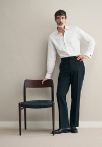 Weißes Hemd mit Knöpfen, schwarze maßgeschneiderte Hose und schwarze Lederschuhe. Hol Stuhl mit dunkler Oberflächenbehandlung und einem schwarzen Lederbezug, auf einem beigen Teppich platziert.