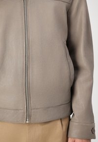 Jaqueta de couro cinza claro com uma textura suave, fecho frontal, bolsos laterais e punhos com botão. Acentuada por elementos de design minimalista.