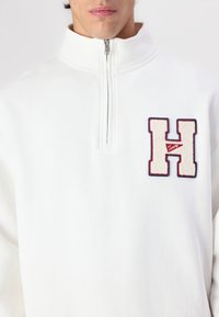Pullover bianco in pile con zip a quarti, con un grande logo in feltro "H" rosso e blu. Texture morbida, colletto alto e zip argento.