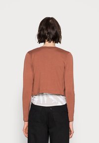 Top corto a maniche lunghe in maglia a coste terracotta con scollatura rotonda, indossato sopra una maglietta bianca tie-dye, abbinato a pantaloni neri.