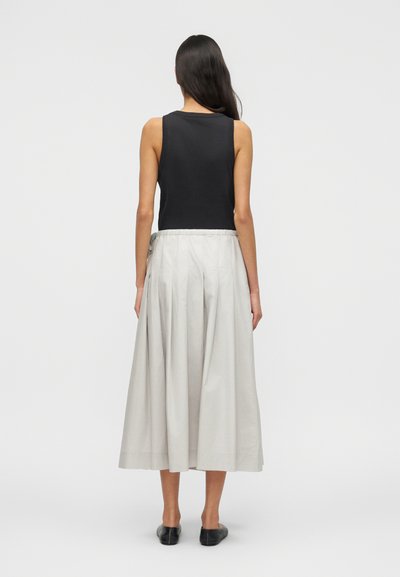 Γυναίκα με μακριά σκούρα μαλλιά, φορώντας μαύρο αμάνικο τοπ, ανοιχτό γκρι φαρδύ παντελόνι και μαύρα επίπεδα παπούτσια, στέκεται σε απλό λευκό φόντο.