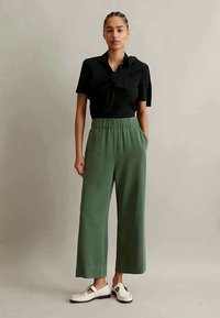 Melns īsām piedurknēm tops ar teksturētu kakla detaļu, saskaņots ar zaļām platajām biksēm un baltām slip-on kurpēm, kurām ir dekoratīvs sprādze.