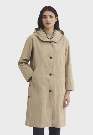 Femme portant un long manteau beige à capuche avec fermeture à boutons et poches latérales, debout devant un fond clair uni.