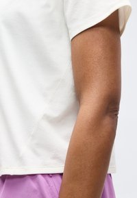 Χέρι και κορμός ατόμου που φοράει λευκό μπλουζάκι με κοντά μανίκια και λιλά παντελόνια, πάνω σε απλό ανοιχτό φόντο.