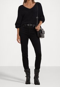 Černý žebrovaný svetr s výstřihem do V, v kombinaci s černými přiléhavými džíny a černými lodičkami na podpatku. Doplňky zahrnují malou texturovanou černou kabelku.