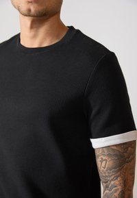 Sort kortærmet skjorte med tekstureret stof og en hvid ribbet kant. Nærbillede viser halsudskæring og detalje af tatovering på overarmen.
