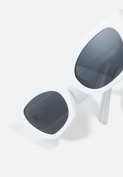 Lunettes de soleil blanches surdimensionnées avec des verres gris foncé, des montures angulaires et des branches épaisses, présentant une finition brillante et une tige transparente.
