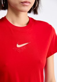 Czerwona bawełniana koszulka z okrągłym dekoltem, z wyciętym logo Nike oraz haftem róży w jaśniejszym odcieniu. Gładka tekstura.