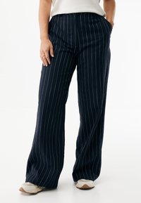 Granatowe spodnie o szerokich nogawkach w białe prążki, z wysokim stanem i bocznymi kieszeniami. Wykonane z teksturowanej tkaniny, zestawione z jasnymi trampkami.