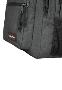 Bolso Eastpak gris con tejido texturizado, múltiples compartimentos, cremalleras negras y un bolsillo lateral de malla. Logotipo de la marca visible en la parte frontal.