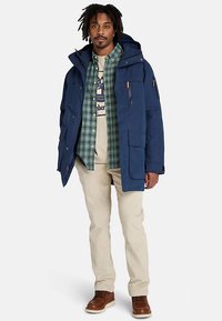 Niebieska wodoodporna kurtka z kapturem, przednimi kieszeniami i zamkami błyskawicznymi. Nosi się ją na zieloną koszulę w kratę i beżowe spodnie, uzupełnione brązowymi butami.