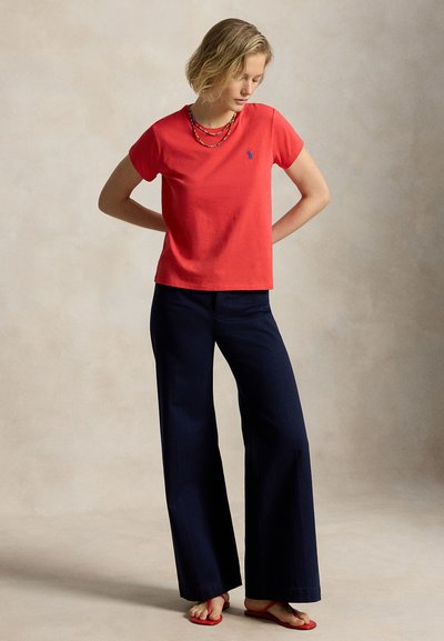 Nő piros rövid ujjú felsőt, sötét bő szárú nadrágot, piros szandált és réteges nyakláncokat visel, háttal áll, kezét a hát mögött tartja, lefelé néz.