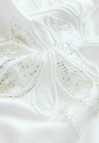 Tissu blanc présentant une broderie florale complexe agrémentée de perles argentées et transparentes, créant une surface texturée et détaillée.