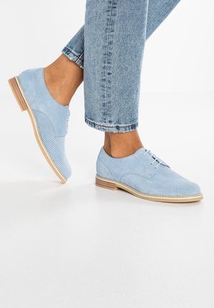 Chaussures Oxford en daim bleu clair avec un design perforé, semelle plate et talon en bois fin couleur tan, associées à un pantalon en jean raccourci.