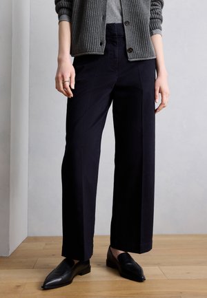 Persoană purtând pantaloni negri cu croială largă, pulover gri și pantofi loafer negri, stând pe podea din lemn, în fața unui perete gri.