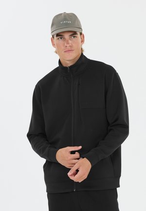 Young man wears a black zip-up jacket and an olive green cap with "VIRTUS" text, standing against a plain white background.