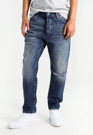 Mann trägt ausgewaschene blaue Jeans mit aufgerissenem Knie und weiße Sneaker, steht vor einem schlichten hellen Hintergrund.