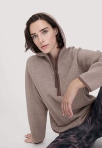 Hellbrauner Fleece-Hoodie mit Reißverschluss, ausgestattet mit einer Kapuze, langen Ärmeln und kontrastierenden dunkelbraunen Akzenten. Modell sitzt mit gemusterten Leggings.