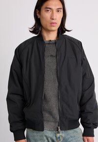 Veste bomber noire avec fermeture éclair à l'avant, poignets et ourlet côtelés, et une poche zippée sur la manche. Portée sur un pull gris texturé.