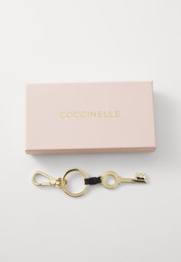 Portachiavi a forma di chiave dorata con anello circolare, cinturino in pelle nera e una scatola rosa marchiata con "Coccinelle" in oro.