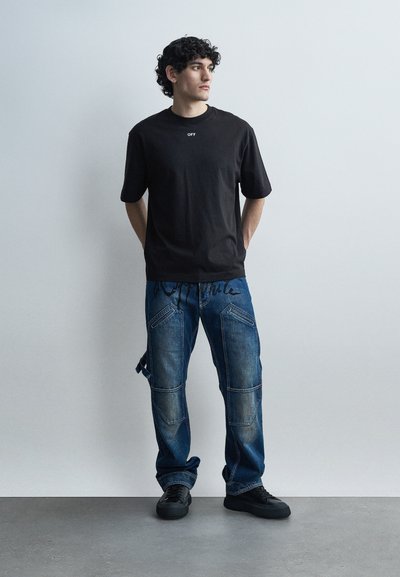 Άνδρας με σγουρά μαλλιά, φορώντας μαύρο μπλουζάκι και μπλε τζιν με μπαλώματα, στέκεται μπροστά σε μια απλή λευκή τοίχο με τα χέρια πίσω από την πλάτη.