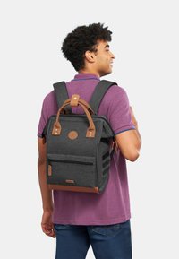 Grauer Stoffrucksack mit Lederakzenten, rechteckiger Form, zwei oberen Traggriffen, verstellbaren Schulterriemen, einer vorderen Reißverschlusstasche und Logoemblem.