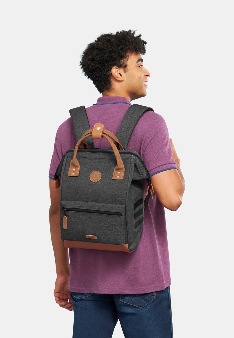 Grauer Stoffrucksack mit Lederakzenten, rechteckiger Form, zwei oberen Traggriffen, verstellbaren Schulterriemen, einer vorderen Reißverschlusstasche und Logoemblem.