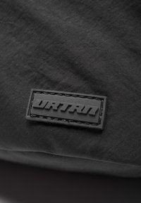Svart tyg med en texturerad yta har en gummihärdad rektangulär etikett präglad med ordet "DRTRN." Stickningen omger etiketten.