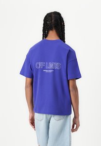 T-shirt oversize violet à manches courtes, avec un texte blanc "OFF LIMITS" et un message en dessous dans le dos. Texture en coton lisse.