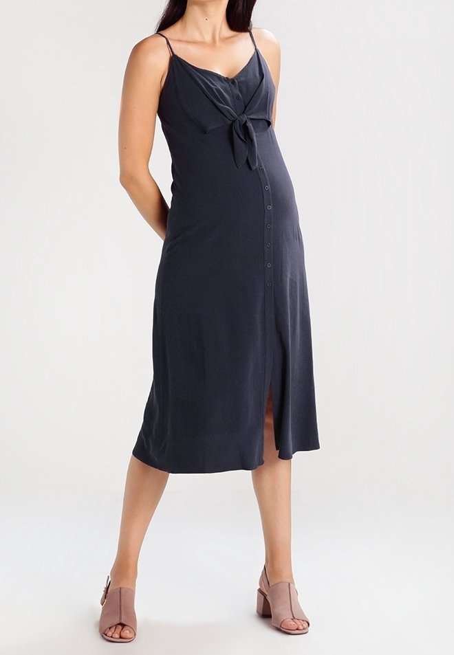 Femme enceinte portant une robe sans manches bleu marine avec des boutons sur le devant et un détail noué, associée à des sandales à talons blocs mauves à bout ouvert.