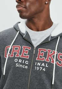 Sudadera con cremallera gris y forro de felpa blanca, con texto rojo y blanco "CRE EKS ORIGINAL Desde 1974." Cuerdas de ajuste en blanco. Parte frontal con cremallera.