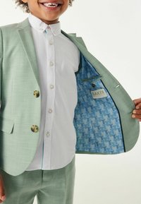 Giacca blazer verde chiaro con bottoni dorati, abbinata a una camicia bianca. L'interno presenta una fodera blu a motivo con un'etichetta.