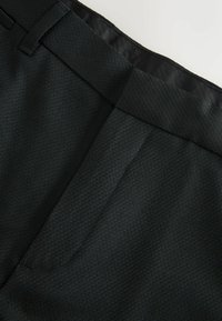 Pantalon vert foncé sur mesure en tissu texturé. Il présente une ceinture lisse et de profondes poches, offrant un design épuré et minimaliste.