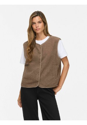 Jeune femme portant un gilet polaire marron boutonné sur un t-shirt blanc et un pantalon noir, debout avec une main dans la poche sur fond blanc.