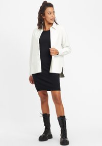 Camicia verde chiaro con bottoni sopra un vestito nero al ginocchio, abbinata a stivali neri con lacci alti fino al ginocchio. Sfondo bianco.