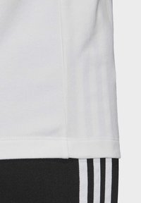 Witte katoenen T-shirt met nette naden en een gladde textuur, gedeeltelijk overlappend met zwarte trainingsbroeken met drie witte strepen.