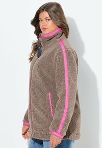 Veste en polaire marron avec des accents roses, col haut, poches latérales et fermeture éclair devant. Texture douce et coupe décontractée.