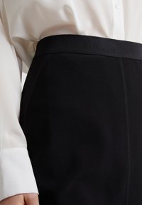 Svart blyertskjol med ribbad midja, slät textur och synliga sömdetaljer. Bärs över en vit, strukturerad blus.