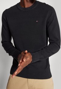 Schwarzer gerippter Strickpullover mit Rundhalsausschnitt, mit einem kleinen Logodetail auf der Brust und engen Bündchen, aus weichem Textilmaterial gefertigt.